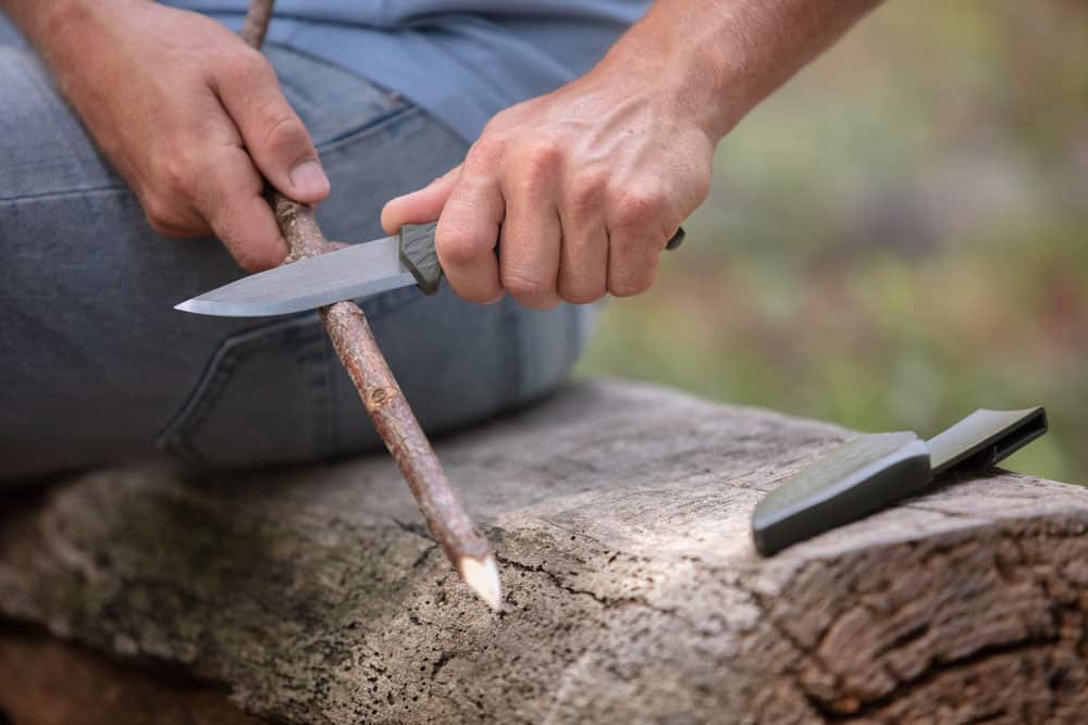 man carving tree branch with fixed blade knife making tinder for fire
