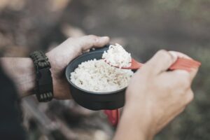 man is eating rice with camping utensils while sitting in the forest