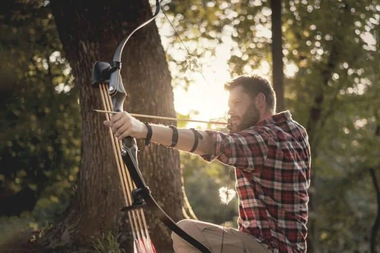 A man kneeling in the woods holding a loaded recurve bow